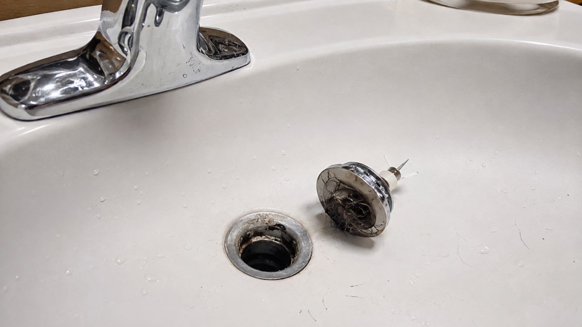 Hair and residue on a sink stopper beside an open bathroom drain