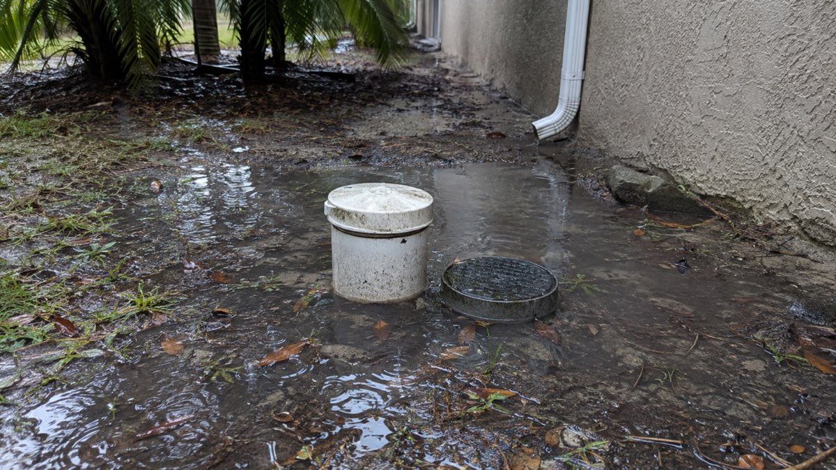 Rainwater pooling around a sewer cleanout and yard drain beside a home